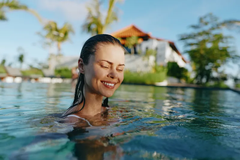 Bright woman enjoying a sunny outdoor pool, smiling with eyes closed, refreshing water, tropical setting, resort vibe, relaxed lifestyle moment, warm breeze