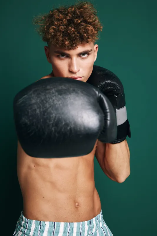 Young male boxer with curly hair wearing black boxing gloves and striped shorts training in front of green background for fitness and combat sport.