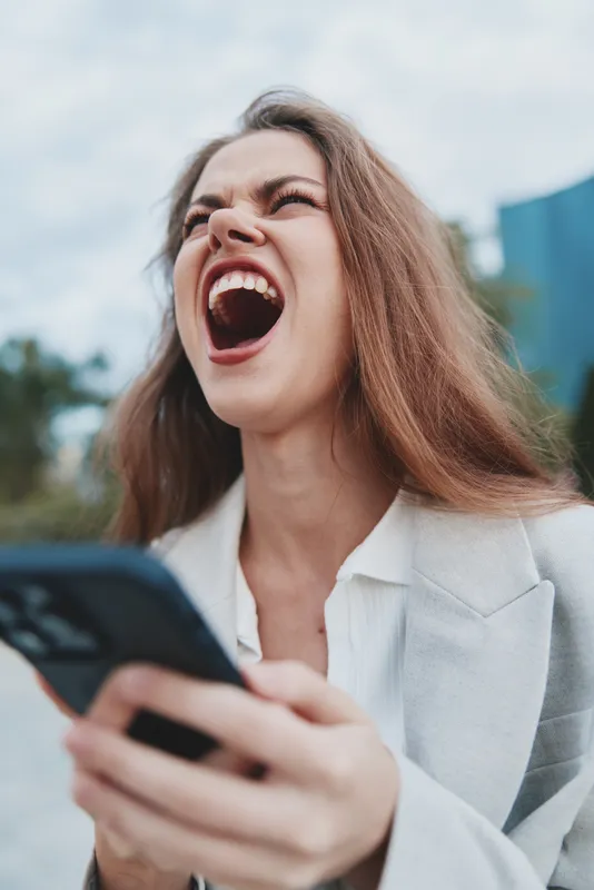 frustrated woman holding smartphone shouting outdoors in casual business attire expressing stress frustration and anger with blurred urban background KEY: frustrated woman, woman, smartphone, stress, 