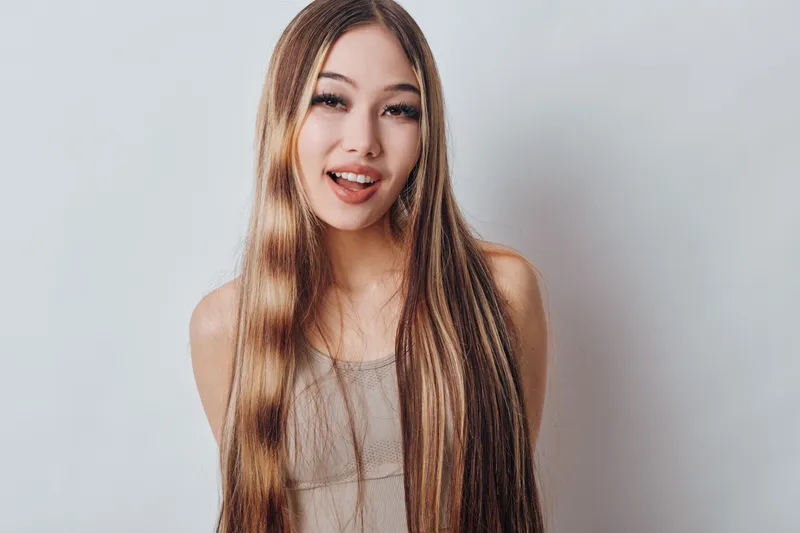 Young woman with long straight hair smiling and posing on white background for beauty portrait in casual wear. Female model with natural makeup and happy expression.