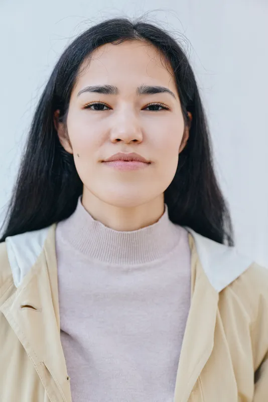 Female with long dark hair and neutral expression, wearing beige jacket over light sweater, standing outdoors against a pale wall, natural daylight, casual fashion mood, relaxed and approachable