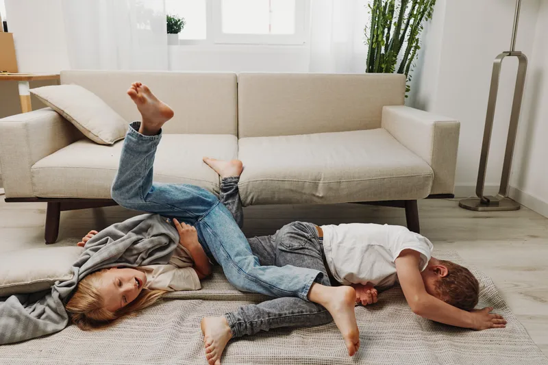 Two boys playfully wrestling on carpet in bright living room with beige sofa and plants, capturing joyful childhood energy and sibling fun.