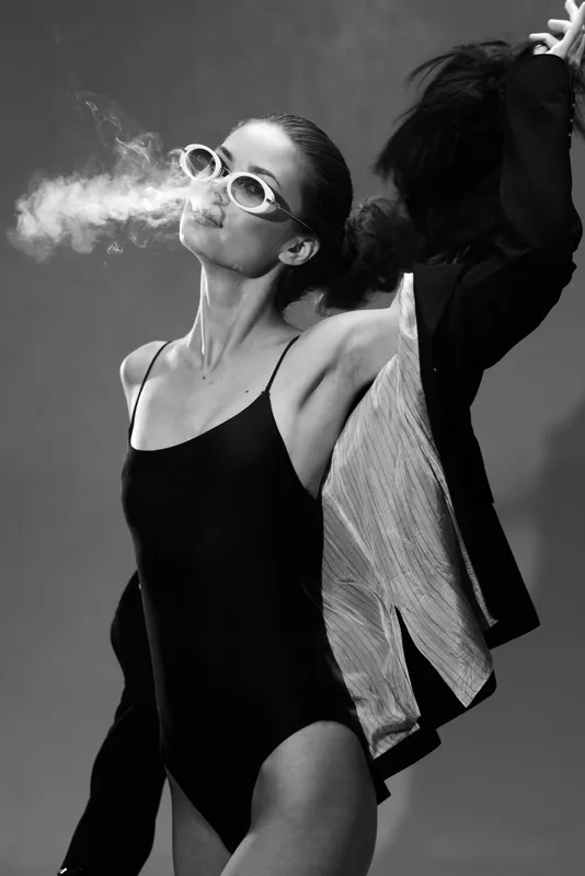 Black and white image of a stylish woman in sunglasses exhaling smoke, wearing a swimsuit and jacket, expressing confidence and cool attitude.