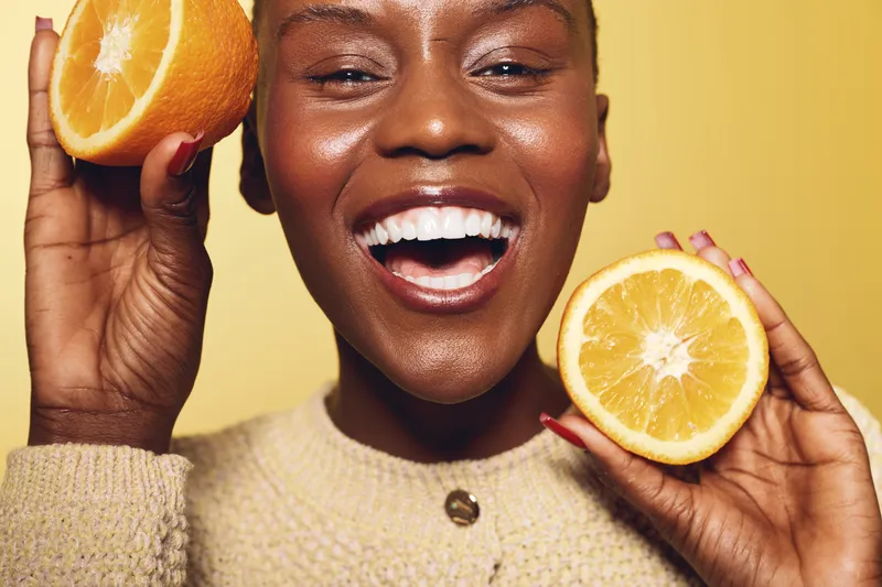 Happy smiling African woman with dark skin holds two halves of fresh orange fruit near face on yellow background. Concept of healthy lifestyle, vitamin C, skincare, and natural beauty.