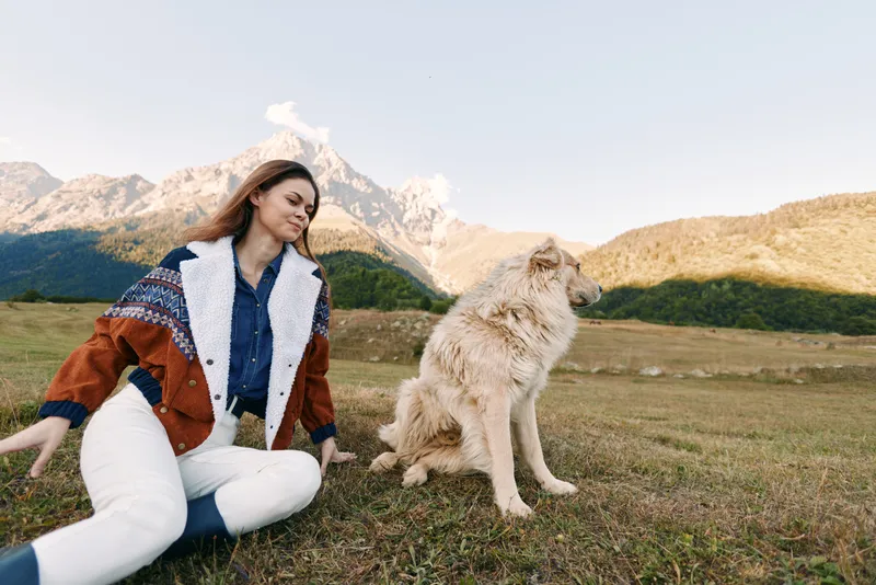 Woman and dog sitting in a mountain meadow outdoors, casual autumn jacket, enjoying nature and pet companionship, relaxed portrait with scenic peaks and open grassy field.