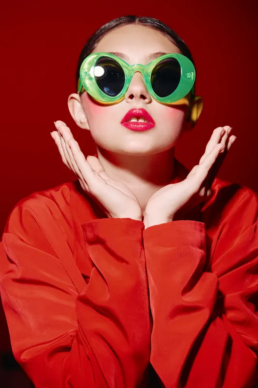 Fashion model in round green sunglasses and bold red lipstick poses with hands on face follower her stylish beauty creative concept on vivid red background.