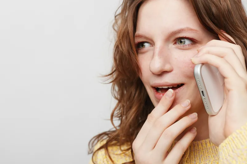 Young woman with curly hair talks on mobile phone, smiling and touching lips. Casual lifestyle, communication, and positive emotions in natural light studio portrait.