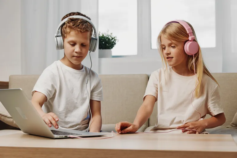 Two children with headphones use a laptop and tablet while sitting on a couch in a bright room. Concept of digital learning, technology, and kids entertainment.