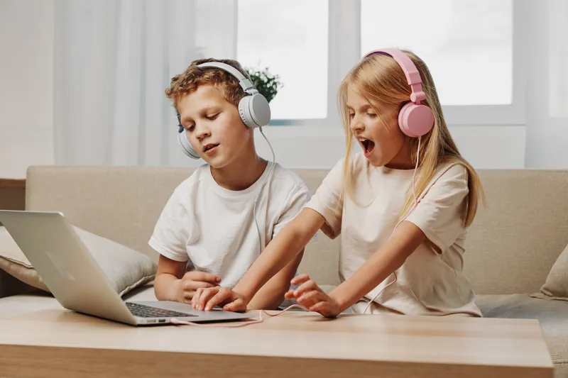 Two children with headphones enjoy music and play together on a laptop at home in a bright living room. Concept of childhood entertainment, technology use, and sibling bonding.