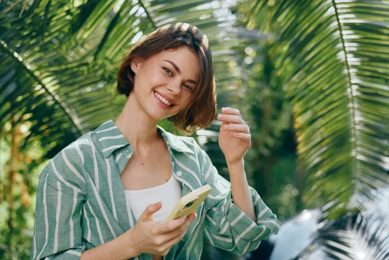 Young woman smiling outdoors, holding a smartphone, framed by lush palm leaves and bright sunlight.
