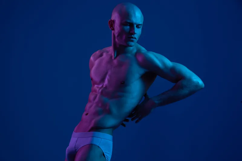 Captivating man posing under vibrant blue and purple lights, exuding confidence and strength in a stylized black and white image that highlights dynamic body lines.