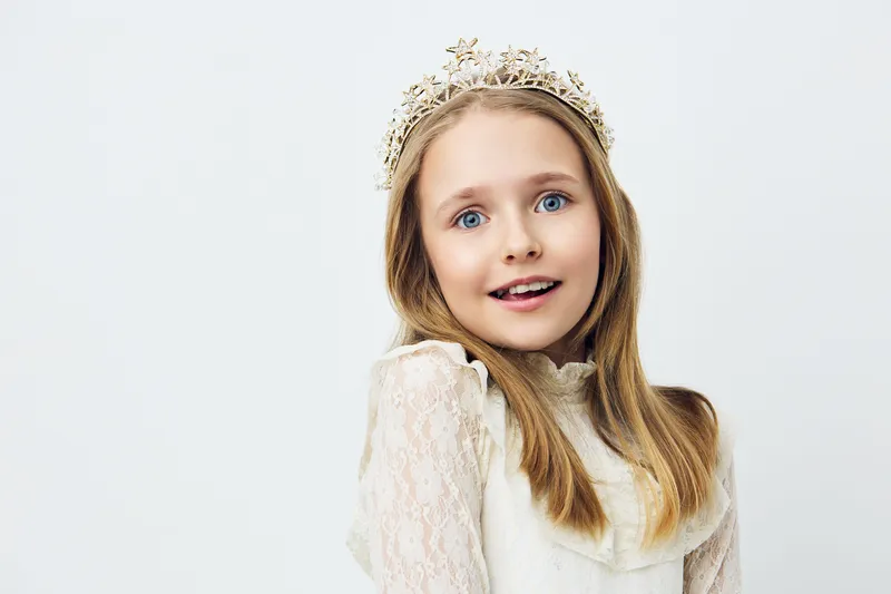 A cheerful young girl wearing a crown and a white dress, smiling brightly against a soft white background, emphasizing innocence and joy in childhood