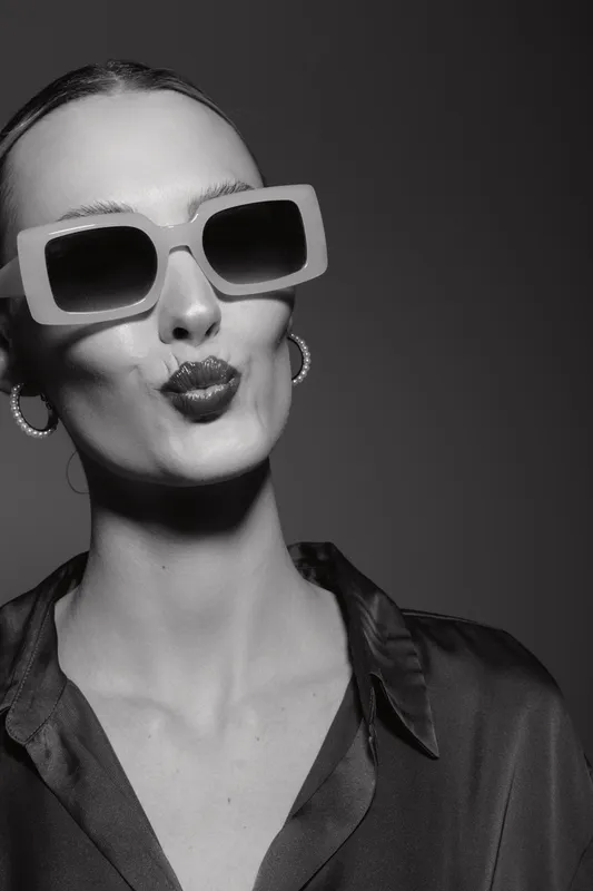 Stylish slender white woman wearing oversized sunglasses and hoop earrings makes a kiss face. Black and white portrait with smooth skin and elegant fashion concept.