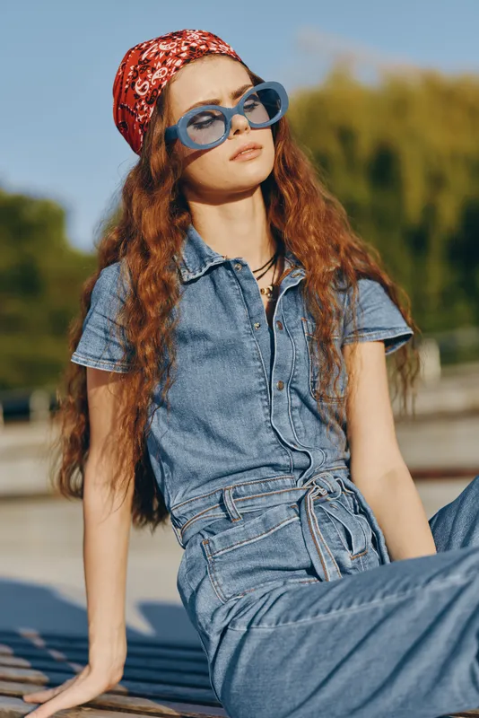 lifestyle woman in modern boho-western denim outfit with red bandana and blue sunglasses, sitting outdoors in warm sunlight, film color tone