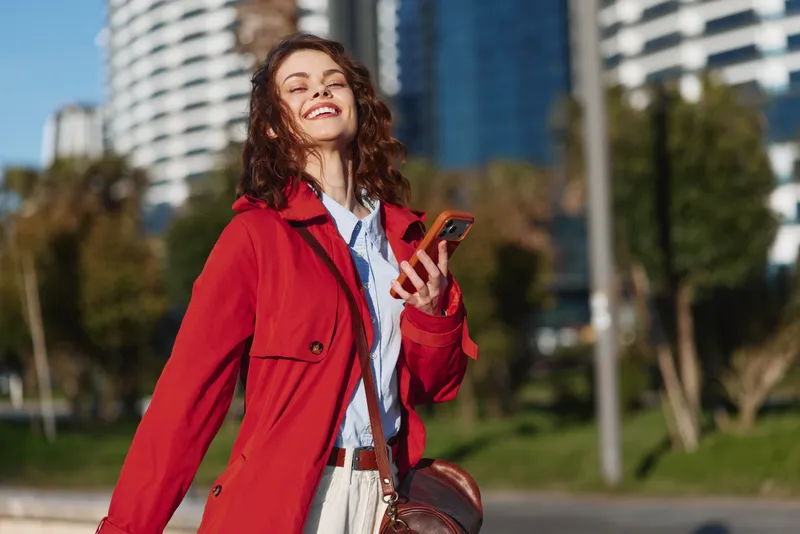 Smiling woman with curly hair wearing red coat holding smartphone outdoors in urban park. Bright sunny day with modern buildings and green trees in background.