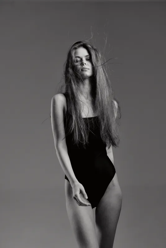 Black and white portrait of a woman with long hair wearing a black bodysuit, posing against a plain background, evoking elegance and simplicity.