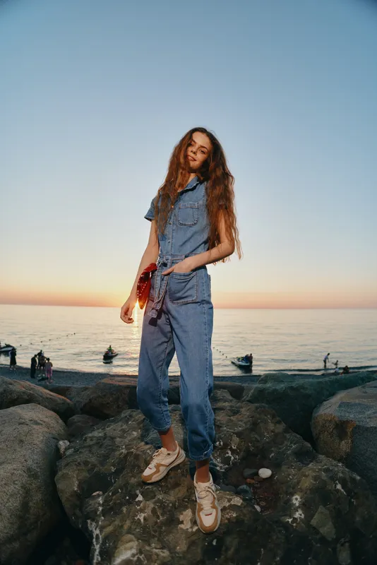 young woman standing on rocky shore at sunset wearing denim outfit with long curly hair and relaxed posture near calm sea and boats