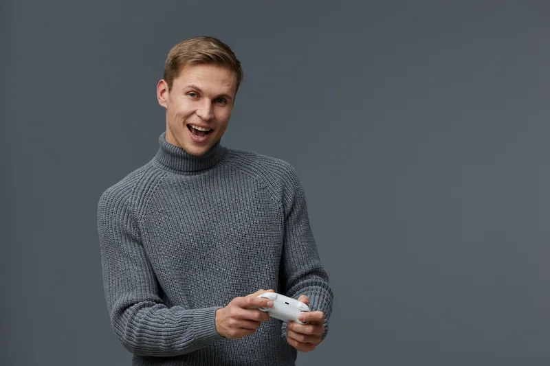 Smiling young man with short brown hair, wearing a cozy gray sweater, holding a gaming console in hand, looking at camera, cheerful expression, indoor studio shot, plain dark background, lifestyle con