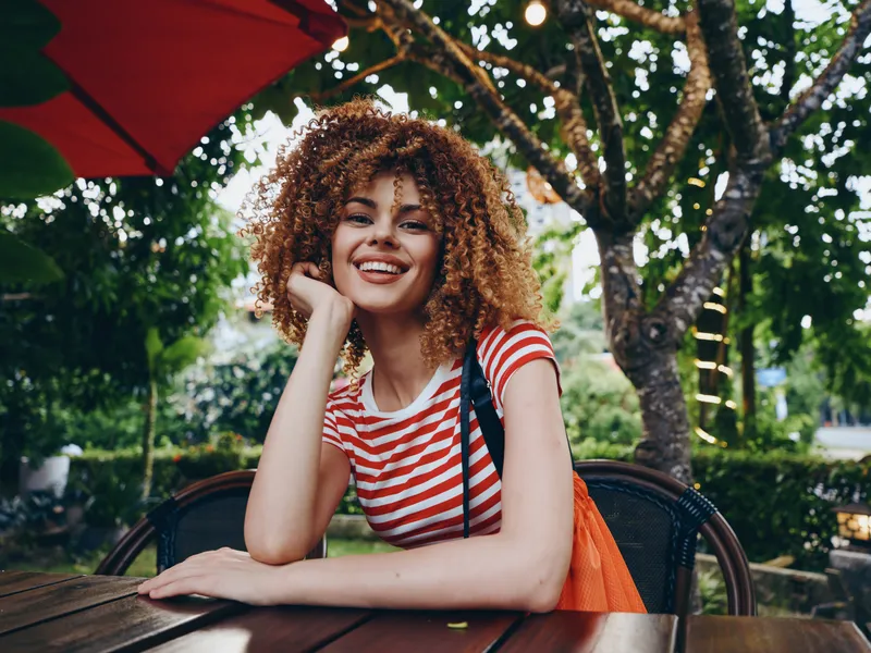 Smiling young woman with curly hair sits at an outdoor cafe table, wearing a red striped shirt and carrying an orange bag, enjoying bright daylight and casual mood.