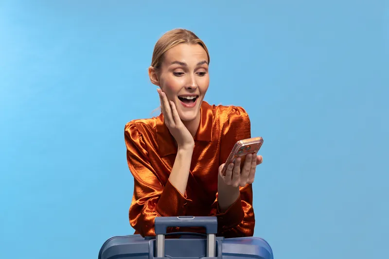 Excited woman in orange blouse looks at smartphone with surprise, sitting behind blue suitcase. Concept for travel, communication, vacation planning, and digital connection with happy expression.