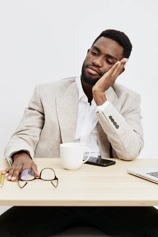 Thoughtful young African American man in beige blazer sitting at desk, resting his head on hand with a pensive expression, surrounded by office supplies, coffee mug, and glasses, isolated on white bac