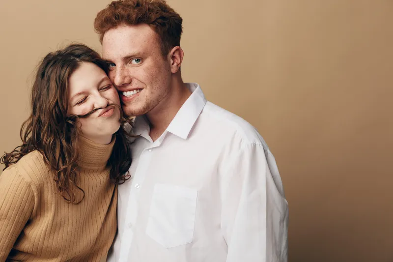 Young couple in casual clothes smiling and posing playfully against beige background. Woman makes mustache with hair, man embraces her with happy and affectionate expression.