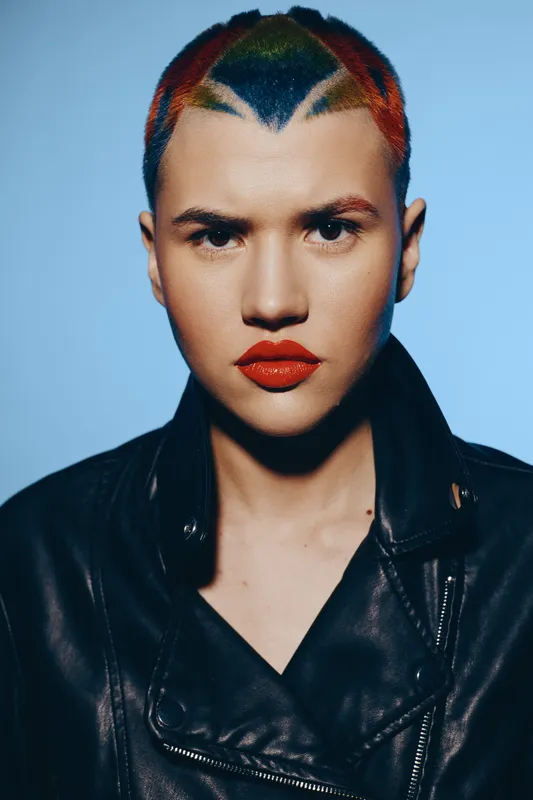 young woman with colorful shaved hair and bold red lipstick wearing a black leather jacket against blue background, intense gaze, modern edgy style portrait