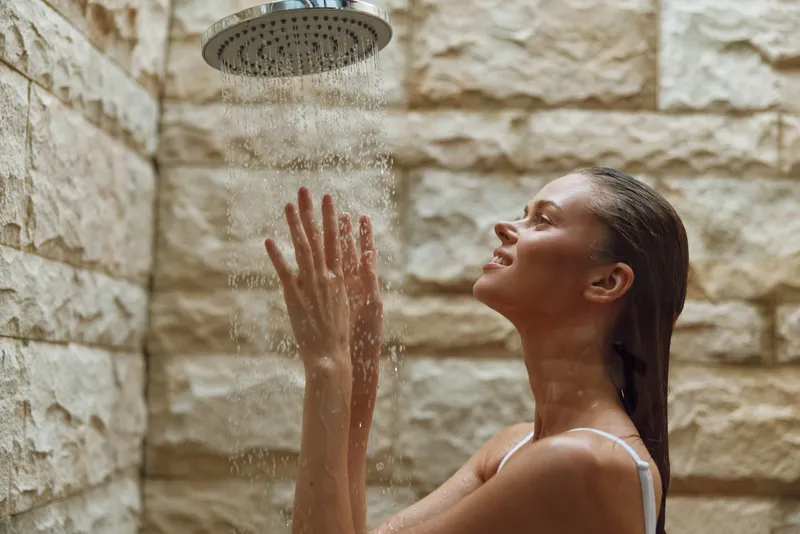 woman showering outdoors with hands raised under rain showerhead, wet hair, serene facial expression, natural stone wall background