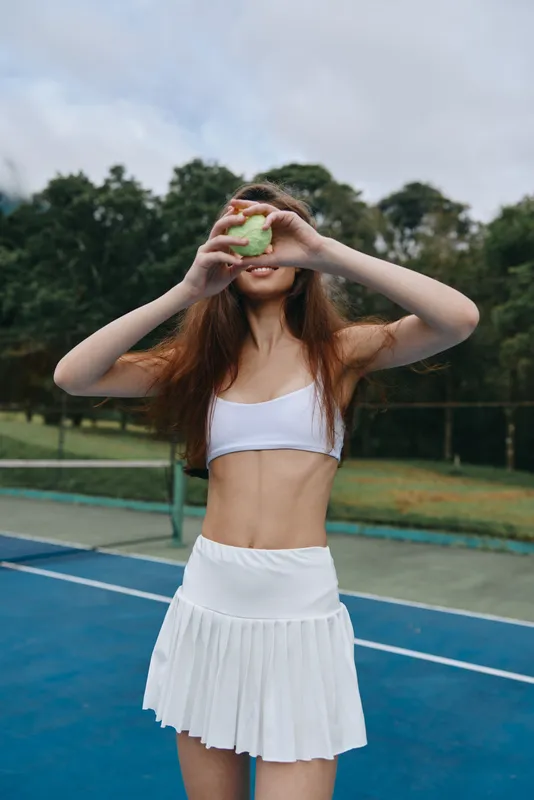 Fit young woman in sporty attire holding a tennis ball on a court, showcasing athleticism and confidence, surrounded by nature in a vibrant, energetic setting