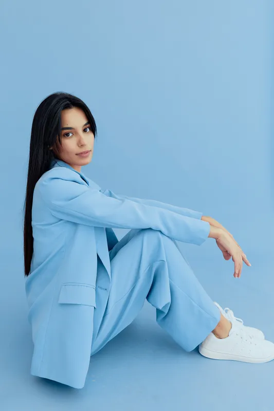 Young woman in light blue suit sitting against blue background, posing with long dark hair and casual white sneakers, calm and stylish atmosphere.