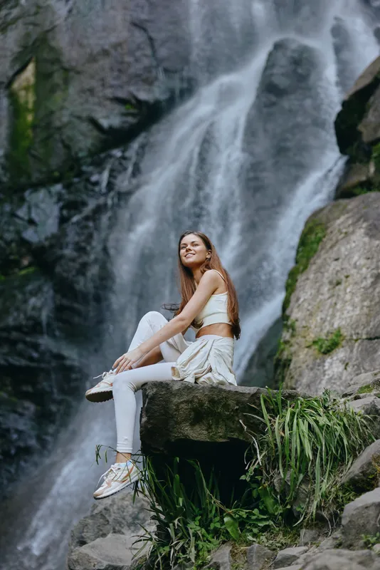young woman sitting near cascading waterfall outdoors wearing casual white clothes, relaxed and enjoying nature with lush green plants around rocks