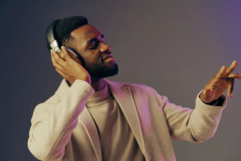 Young Black man enjoying music with headphones, dressed in stylish beige coat against a gradient background, expressing joy and relaxation through his gestures