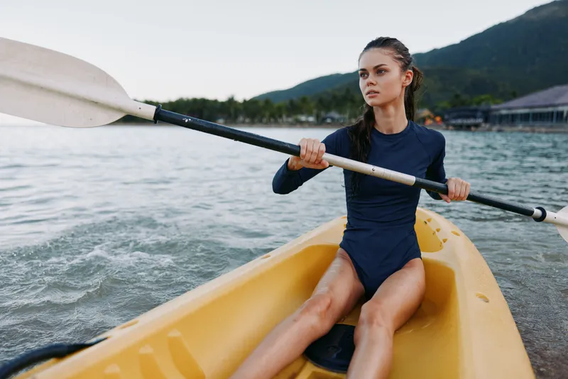 A focused young woman paddles a yellow kayak on calm water, gripping the paddle with strength and balance as mountains rise in the background