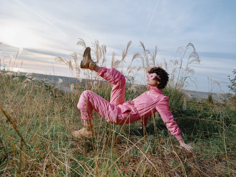 Outdoor scene with a person in a pink jumpsuit lounging on tall grass on a hillside, boots kicked up, relaxed pose, warm sunlight highlighting natural fashion in open countryside.