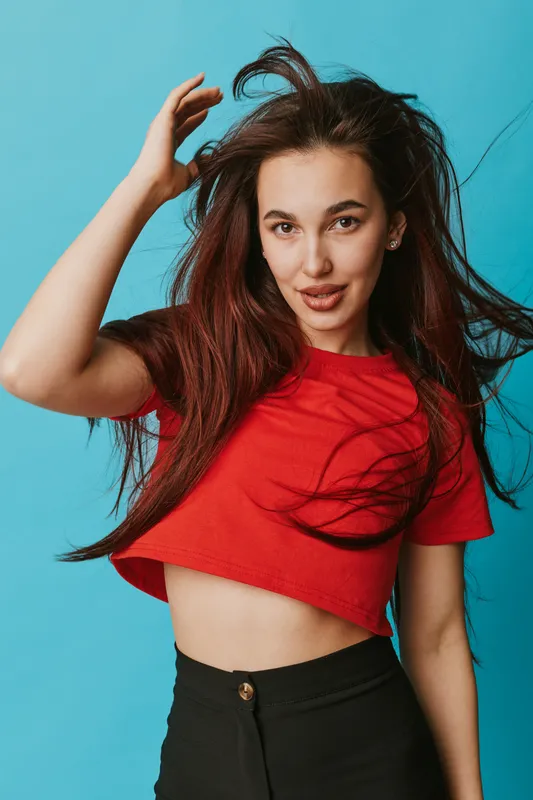 Young woman with long hair wearing red crop top and black pants posing against blue background smiling and touching hair in casual fashion portrait studio shot.