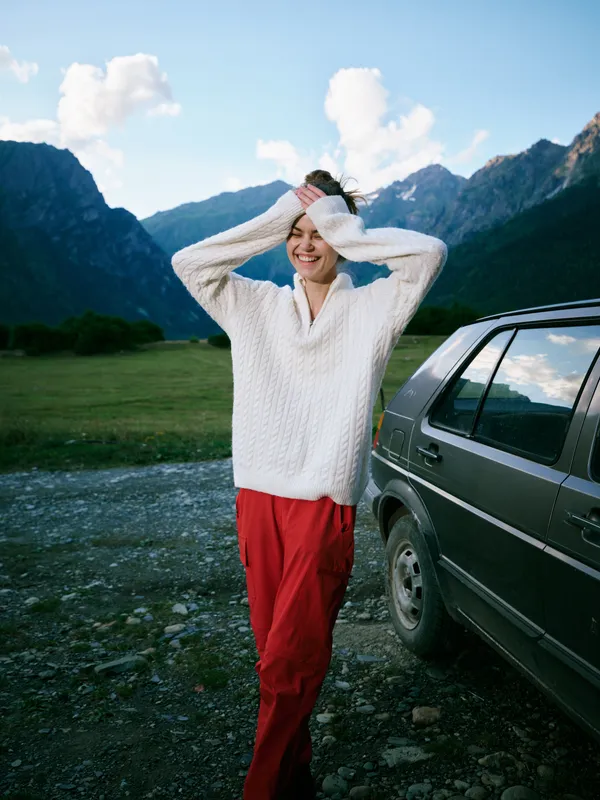 Outdoor scene with a smiling woman beside a parked car in a mountain valley, wearing a white sweater and red pants, enjoying fresh air and wide alpine scenery