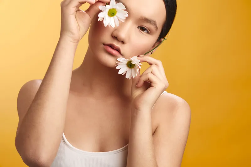 asian woman holds daisies near her face in a studio portrait with a warm yellow backdrop, conveying softness, calm, and natural beauty.