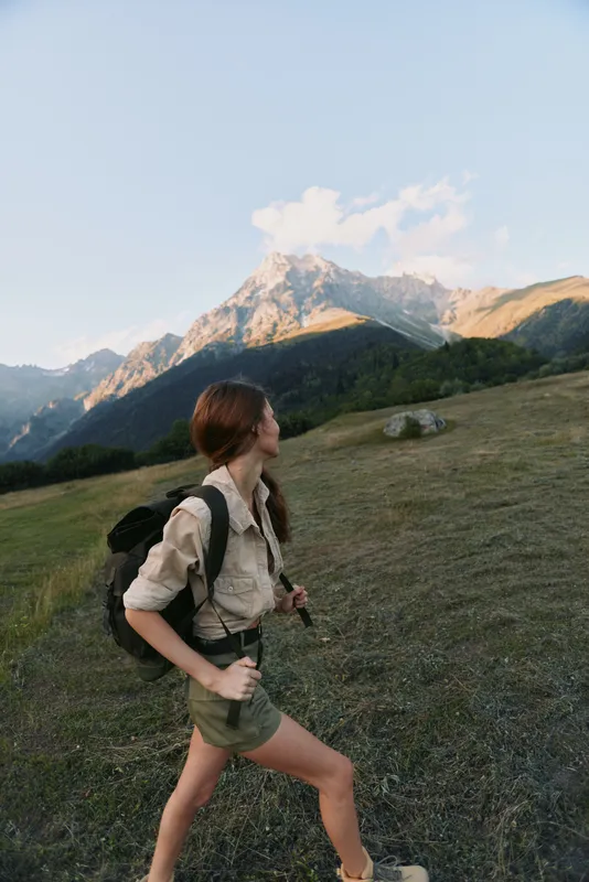 Hiking woman with a backpack strides through a grassy meadow toward distant mountains, focused and determined in a natural outdoor setting, her hair tied back, soft light highlighting the landscape