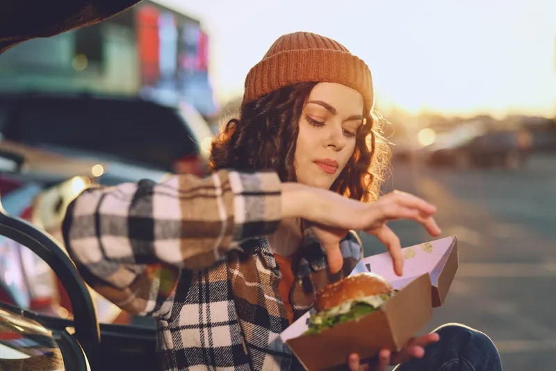 Woman in beanie eats burger from takeout box in urban sunset, candid lifestyle portrait with golden hour glow, authenticity and mindful living captured in emotional storytelling moment.