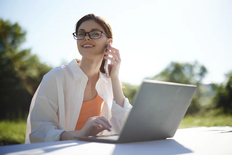 Young woman in casual wear working on a laptop outdoors, enjoying the sunny day while smiling and interacting on the phone