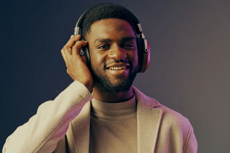 Young African man enjoying music with headphones, dressed in stylish attire, smiling against a colorful gradient background that evokes joy and relaxation