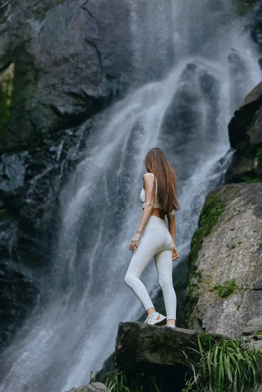 Woman in white activewear stands confidently on rock by a cascading waterfall surrounded by lush greenery in a serene natural setting during daytime.
