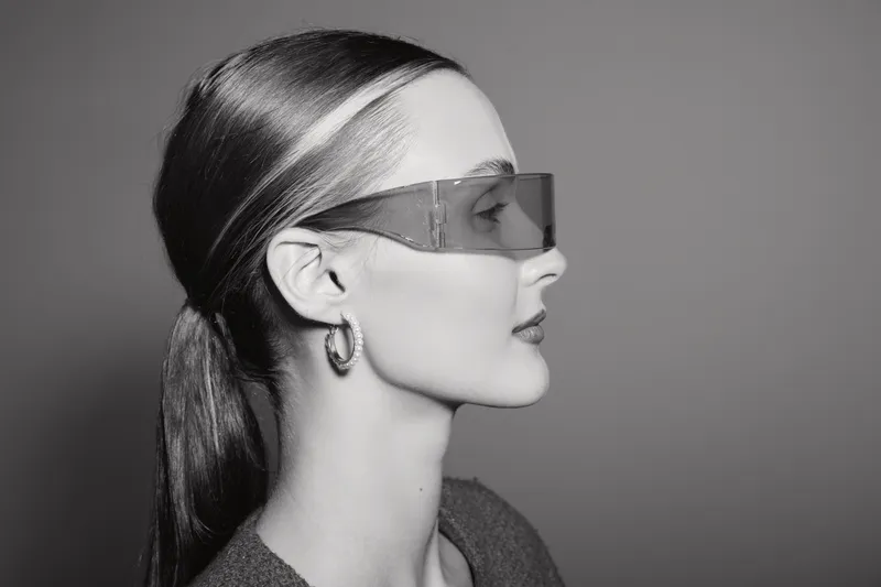 Profile of a young white woman with light skin wearing transparent sunglasses and hoop earrings. Minimalistic black and white portrait with sleek hairstyle.