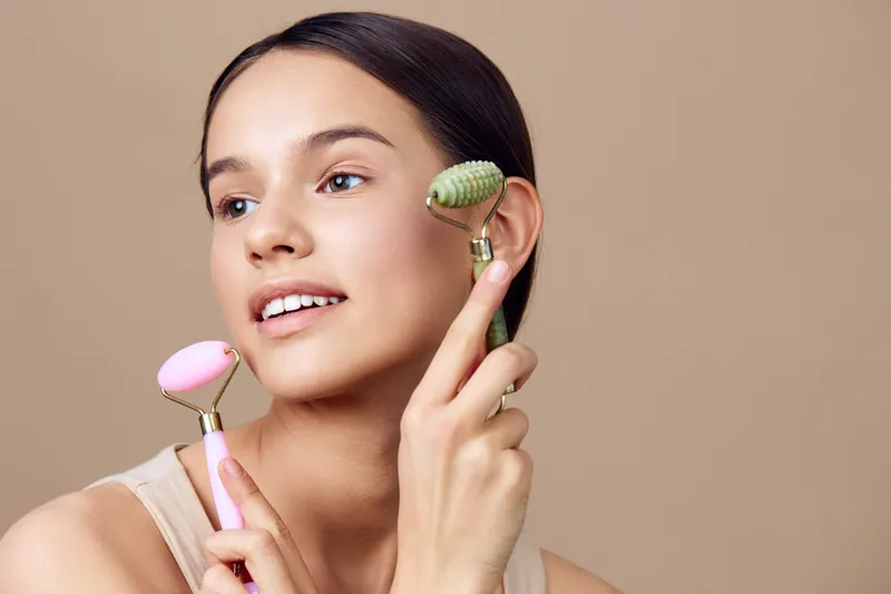 Young woman holding facial rollers in pastel colors, promoting beauty and skincare, joyful expression, soft beige background, seasonal wellness concept