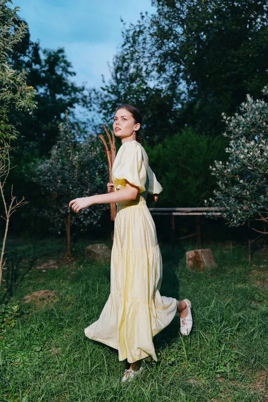 Young woman in a flowing yellow dress walks through a lush garden, capturing a serene outdoor moment with soft light, movement and natural elegance.