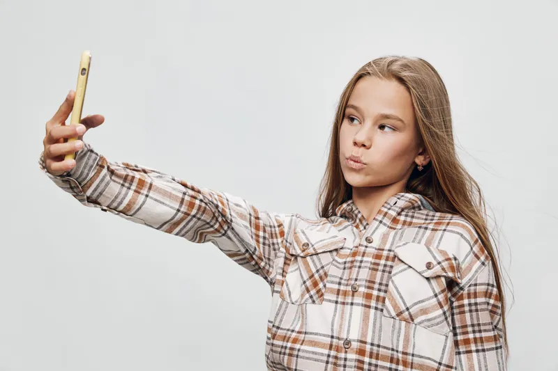 Teenage girl in plaid shirt taking selfie with smartphone, posing with pout lips and long hair in casual studio setting on white background, youth and social media concept.