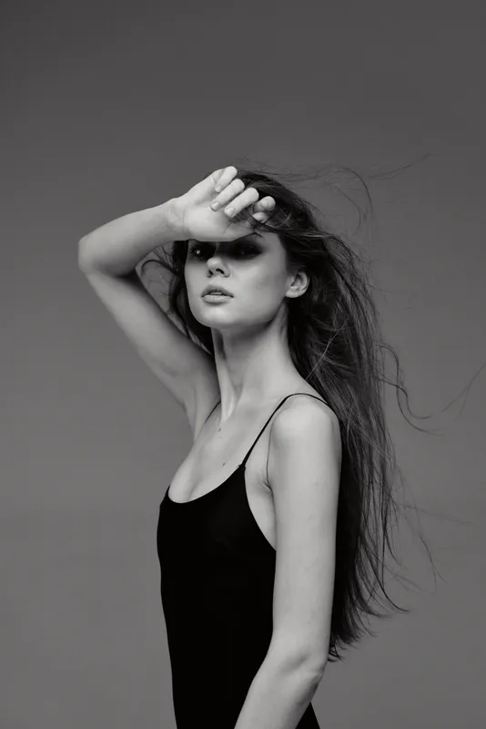Black and white portrait of a young woman with long hair wearing a sleeveless top and covering her forehead with her hand.