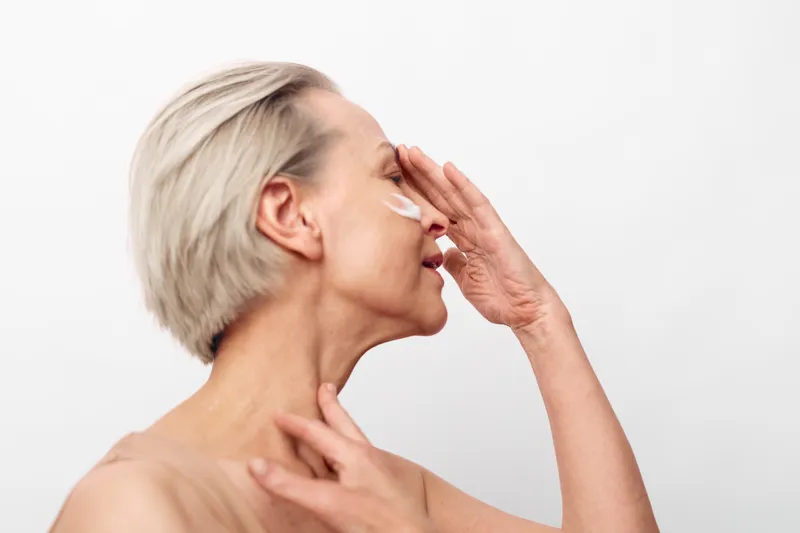 Mature woman with short blonde hair applying cream on face and neck, eyes closed, isolated on white background. Concept of skincare, self-care, and anti-aging beauty routine.