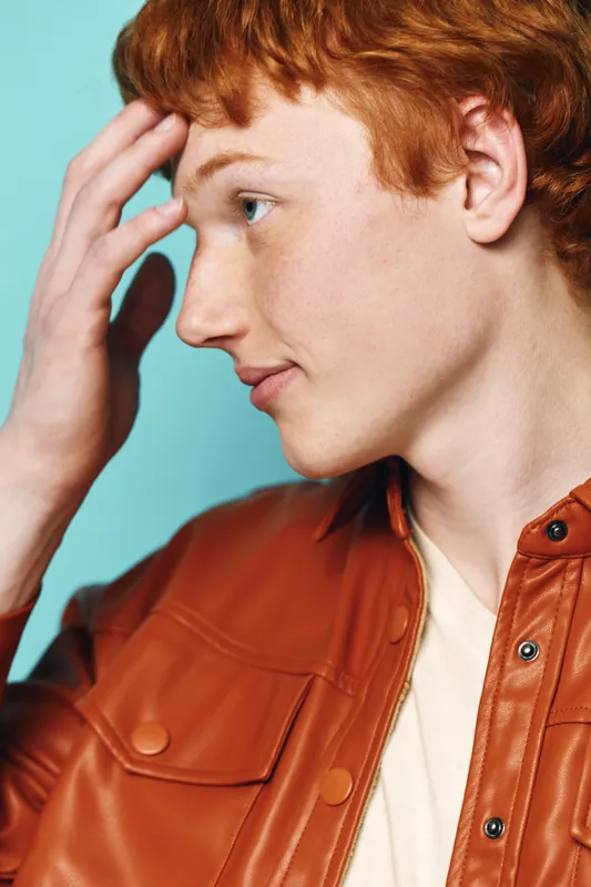 Portrait of young redhead man with hand on forehead wearing brown jacket and white shirt on blue background. Casual style, thoughtful expression, side profile closeup with clean skin and freckles.