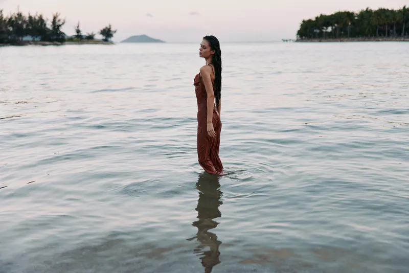 A lone woman in a flowing red dress stands waist-deep in calm coastal water, her reflection shimmering as gentle waves lap the shore and create a tranquil, cinematic mood.